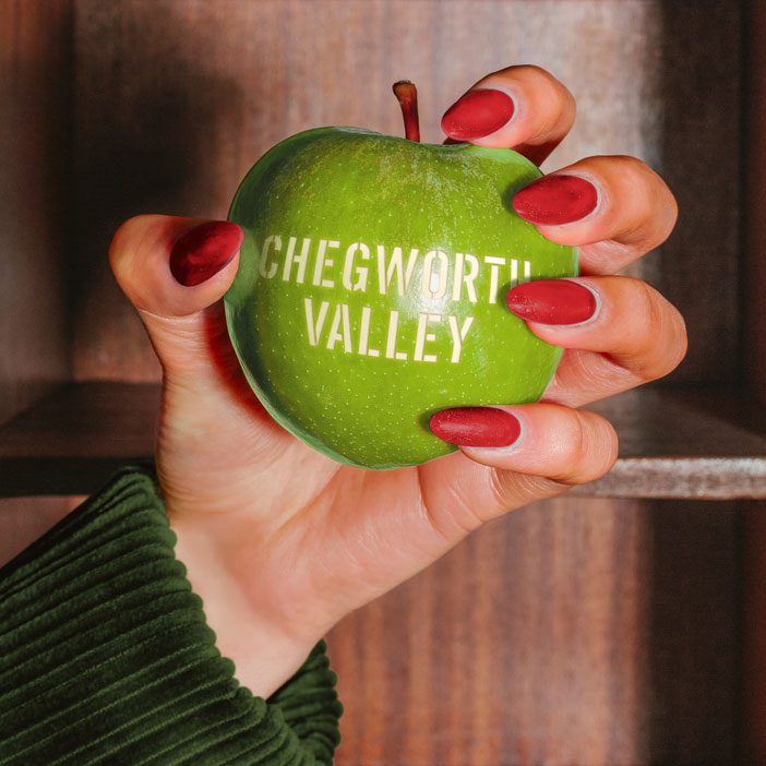 Hand holding a green apple stamped with Chegworth Valley branding, set against rustic wooden shelves, highlighting fresh orchard produce and natural juice heritage.