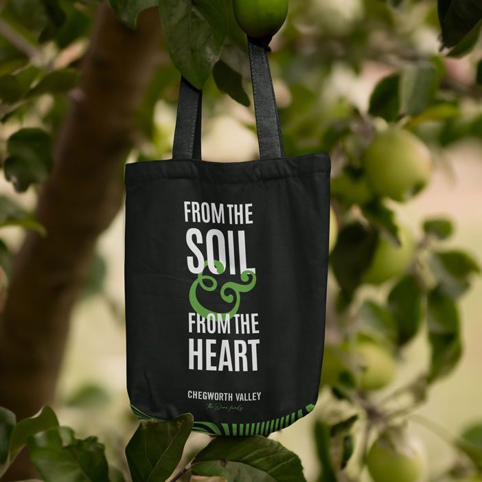 Chegworth Valley branded tote bag hanging in an apple tree, featuring the slogan “From the Soil & From the Heart” in a British apple orchard.