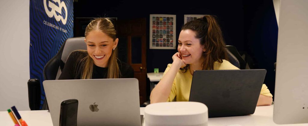 Ellie and Caroline smiling while working together on laptops at a desk in a modern creative office.